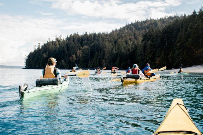 The image shows a group of people kayaking in a body of water. The kayakers are spread out, with some closer to the camera than others. They are surrounded by lush green trees and a cloudy sky. The water is calm, and the overall scene is peaceful and serene. The kayaks are of different colors, including yellow and green.