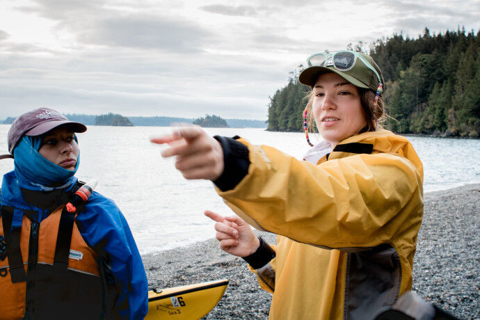 The image shows two people on a rocky beach near a body of water with islands in the background. The person on the right is wearing a yellow raincoat and a baseball cap and is pointing towards something in the distance. The person on the left is wearing a blue jacket and a red baseball cap and is looking towards the same direction. A yellow kayak is visible near the bottom of the image.