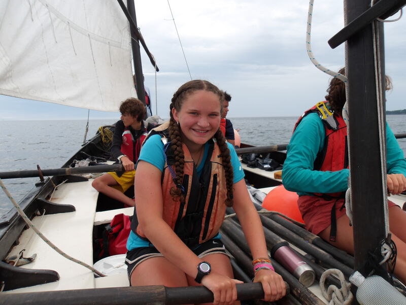 The image shows a young girl with braided hair wearing a life vest, smiling while sitting in a sailboat. She appears to be holding oars. Other people are visible in the boat, also wearing life vests. The background shows water and a cloudy sky.