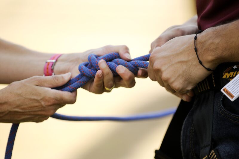 The image shows two people's hands working with a blue rope. One person is holding the rope, while the other is tying a knot. The focus is on the knot-tying process, suggesting an activity like climbing or sailing. The background is blurred, emphasizing the hands and the rope.