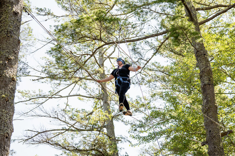 The image shows a person walking on a tightrope course in a forest. The person is wearing a helmet and safety harness. They are carefully balancing on a rope bridge suspended between trees. The background is filled with green foliage and sunlight.