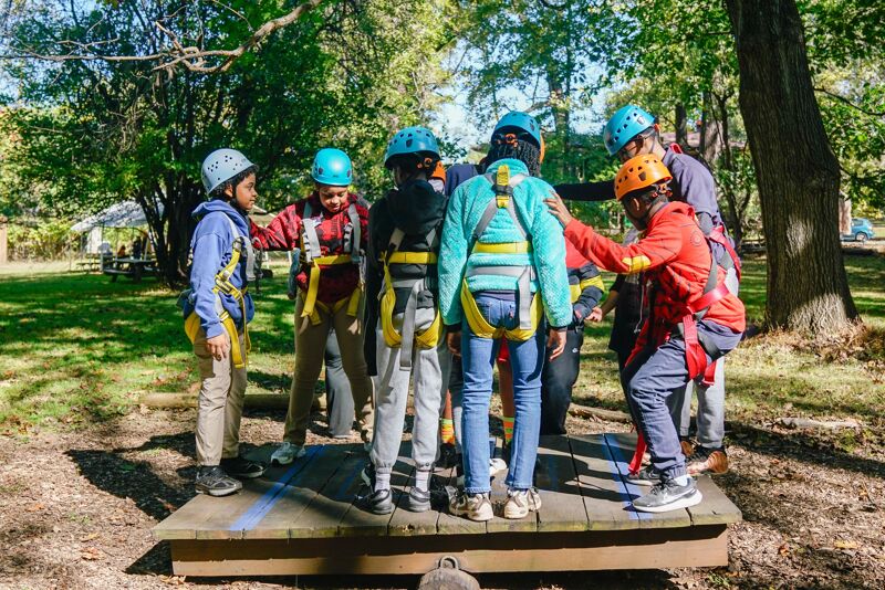A group of young people wearing helmets and harnesses are gathered on a wooden platform in a wooded area. They appear to be participating in some kind of outdoor adventure activity, possibly a ropes course or team-building exercise. The participants are interacting with each other, some are helping to adjust the harnesses of others.