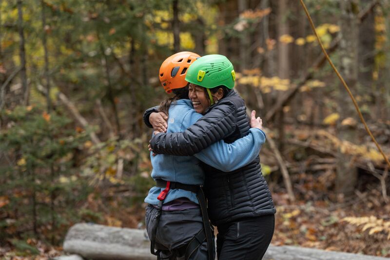 The image shows two people wearing helmets embracing each other in a forest setting. They appear to be celebrating or comforting each other. The person on the left is wearing an orange helmet, while the person on the right is wearing a green helmet. The background is filled with trees and foliage.