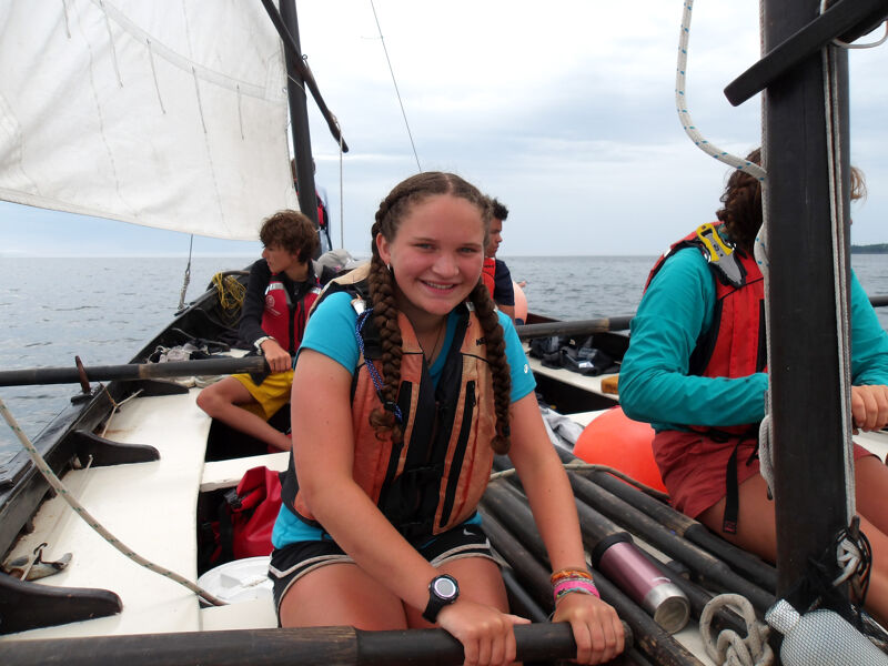 A young woman in a life jacket smiles while sitting on a sailboat. She is surrounded by other people, also wearing life jackets, and sailing equipment. The boat is on the water, and there is a sail visible in the background. The sky is overcast.