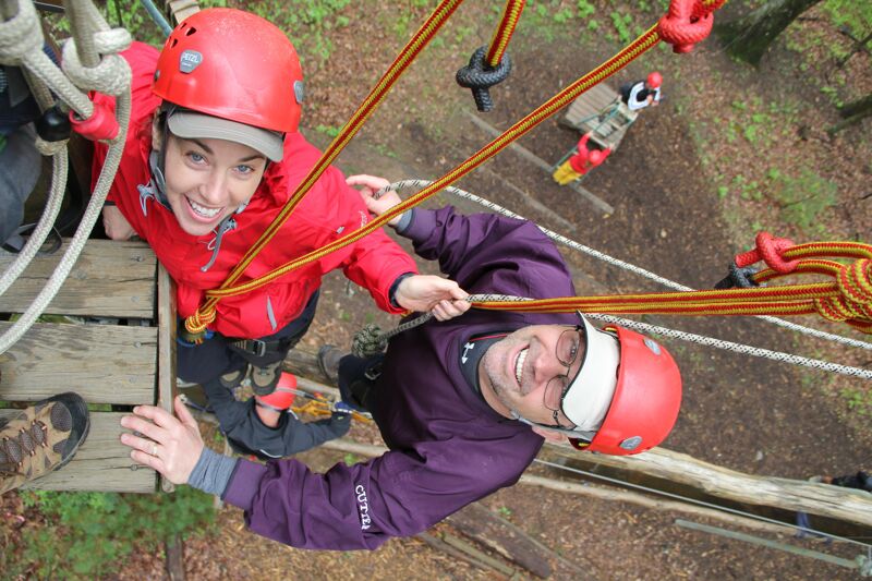 From a high-angle perspective, the image captures two individuals on a ropes course, secured with helmets and harnesses. The woman, wearing a red jacket, smiles at the camera, while the man in a purple shirt looks upwards. Ropes and wooden platforms form the intricate course structure, set against a backdrop of trees.
