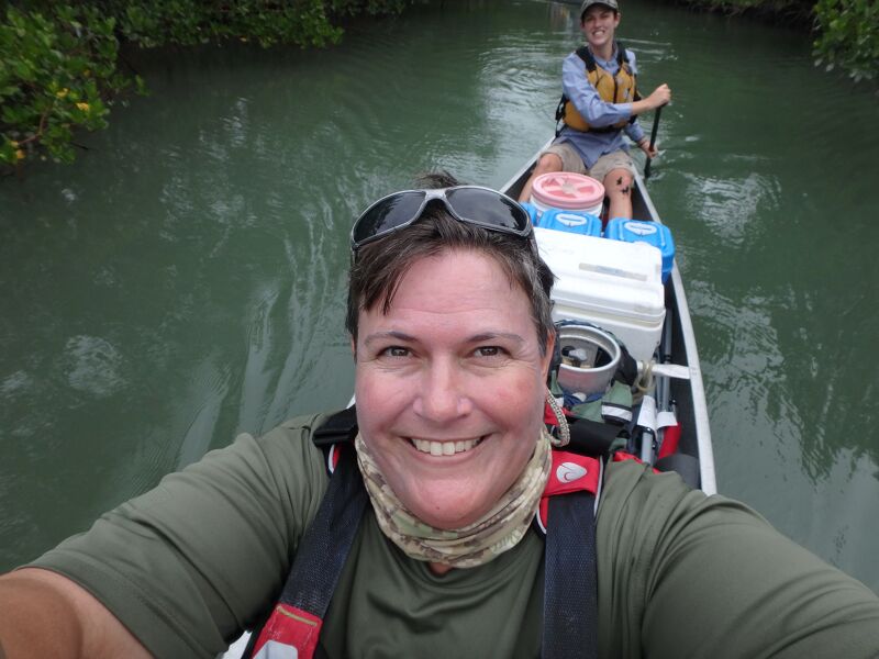 A selfie shows a smiling woman in the foreground, wearing sunglasses on her head and a neck gaiter. She's in a canoe loaded with gear, including a cooler and other supplies. Behind her, another person is paddling the canoe through a narrow waterway surrounded by lush green vegetation. The overall scene suggests an outdoor adventure or expedition.