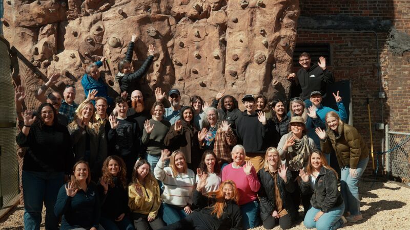 A group of approximately 30 people are posing for a photo in front of a rock climbing wall. Most of them are smiling and waving. The group appears to be diverse in age and ethnicity. The photo is taken outdoors, with a brick building visible in the background. The overall impression is one of camaraderie and teamwork.