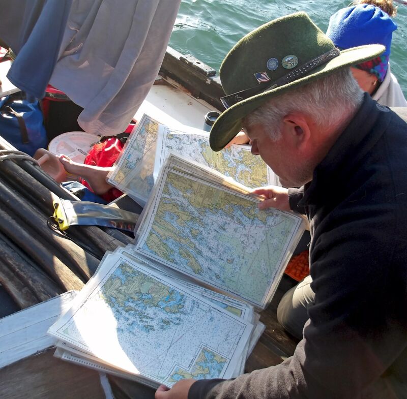 A man wearing a green hat is intently studying a stack of maps. He is seated in what appears to be a boat, surrounded by other people and gear. The maps are spread out, suggesting he is navigating or planning a route. The scene conveys a sense of adventure and exploration, with the man focused on the task at hand.