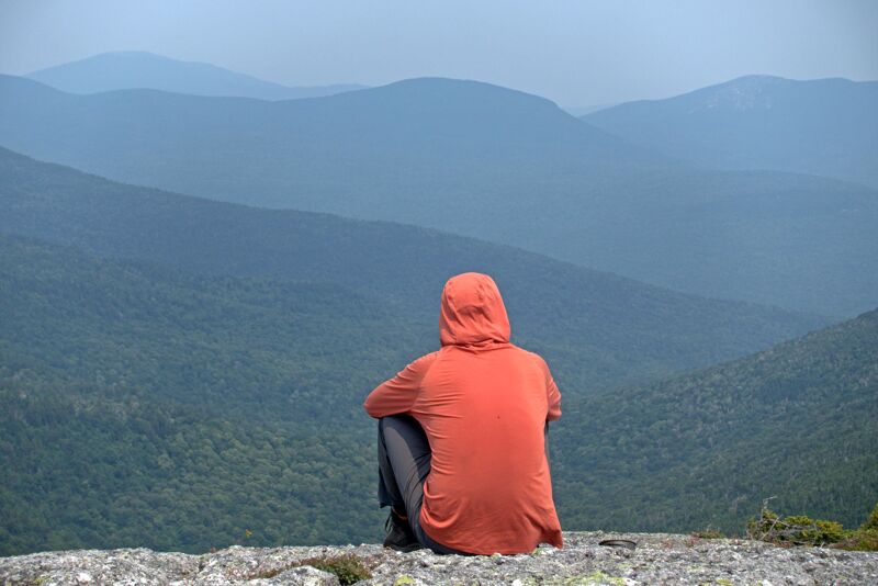A person wearing an orange hooded jacket sits on a rocky outcrop, gazing out at a vast landscape of rolling, forested hills. The hills fade into a hazy blue distance, creating a sense of depth and tranquility. The person is positioned with their back to the viewer, emphasizing the solitary nature of the scene and inviting contemplation of the expansive view.