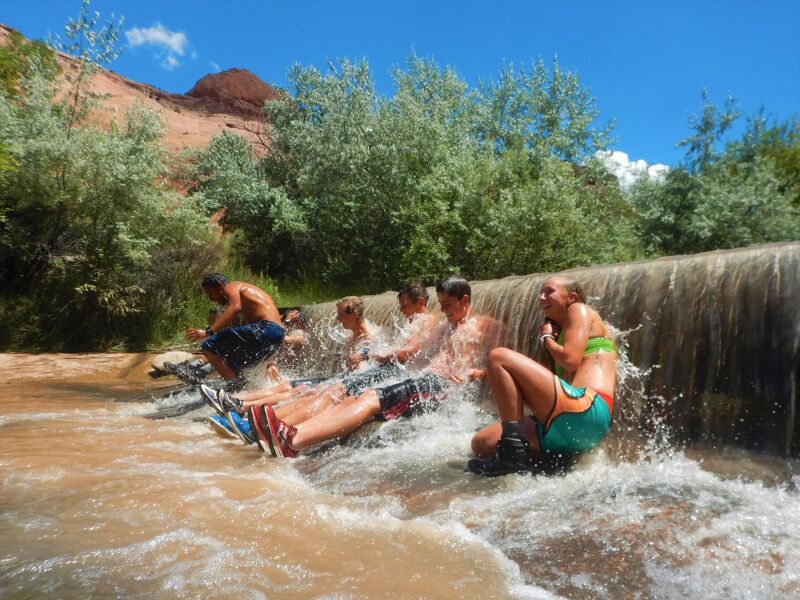 A group of people are sitting at the top of a small waterfall, enjoying the water cascading over them. They appear to be having fun, with smiles and laughter evident. The scene is set in a natural environment, with lush greenery and a clear blue sky in the background. The water is flowing rapidly, creating a refreshing and playful atmosphere.
