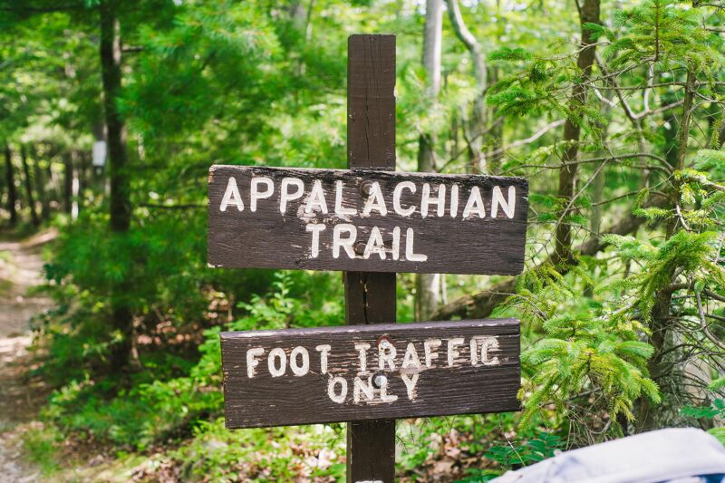 The image shows a wooden signpost in a wooded area. The top sign reads "Appalachian Trail" in white letters on a dark background. Below it, another sign says "Foot Traffic Only" in a similar style. The signs are weathered, and the surrounding foliage is lush and green. A trail is visible in the background, suggesting the start or continuation of the Appalachian Trail.