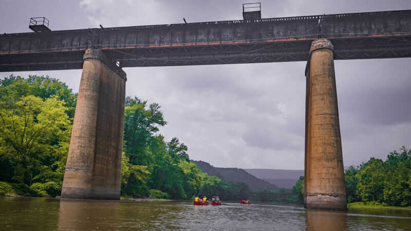 The image shows a low-angle view of a long, weathered bridge supported by large, cylindrical pillars over a body of water. Several small boats with people are on the water, heading towards the bridge. Lush green trees line the banks of the water, and a distant mountain range is visible under a cloudy sky.