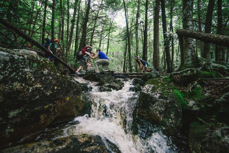 A group of hikers carefully crosses a rocky stream in a dense forest. The water rushes over the rocks, creating small waterfalls. The hikers are using logs and rocks to navigate the stream. The surrounding forest is lush and green, with tall trees and moss-covered rocks. The scene conveys a sense of adventure and the beauty of nature.