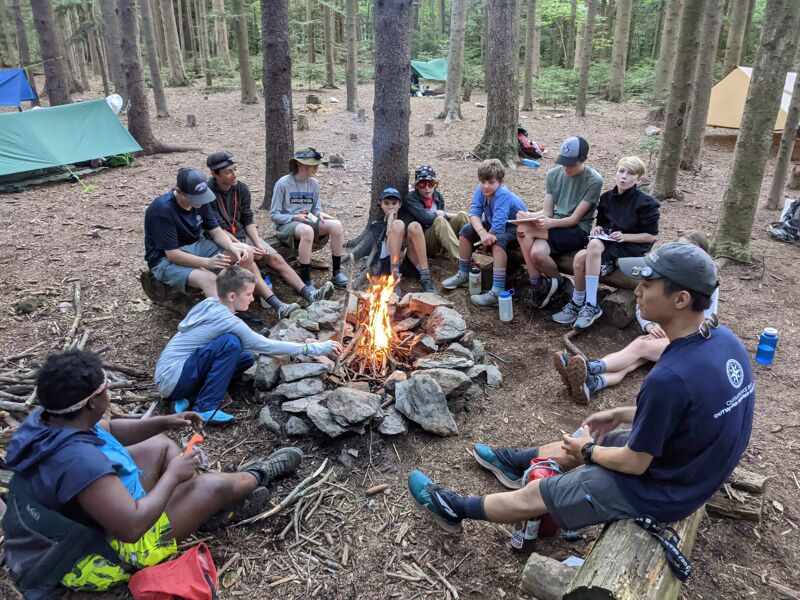 A group of young people are gathered around a campfire in a wooded area. They are sitting on logs and rocks, and some are talking while others are looking at the fire. There are tents pitched in the background, suggesting they are camping. The atmosphere seems relaxed and communal, indicative of a group activity like a scout trip or summer camp.