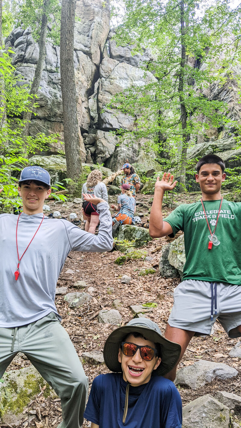 The image shows a group of people on a rocky, wooded trail. In the foreground, three young men are posing for the camera. One is wearing a hat and sunglasses, while the other two are wearing caps and t-shirts. In the background, other people are visible further up the trail, surrounded by trees and rocks.