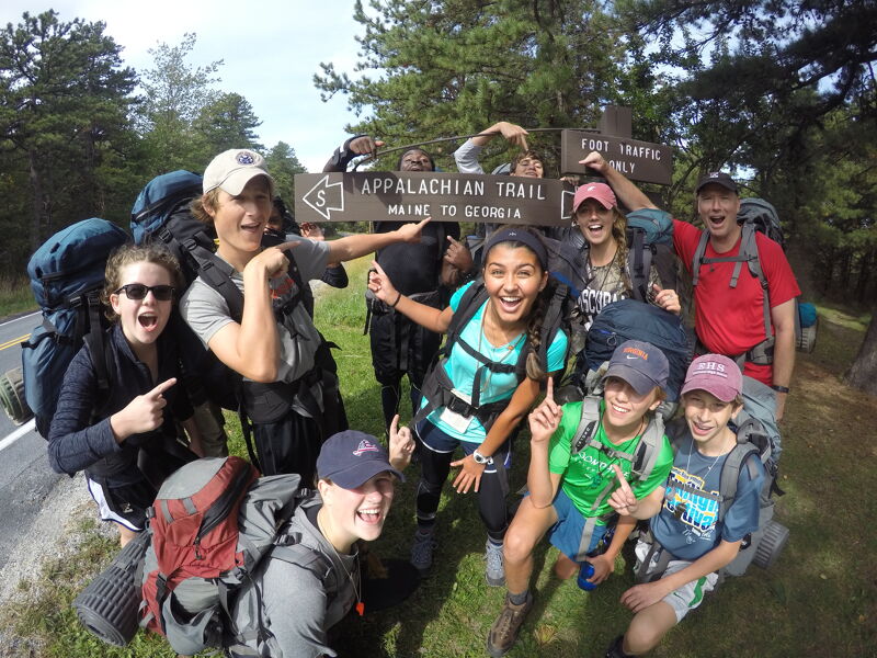 A group of young hikers poses near an Appalachian Trail sign. They are wearing backpacks and hiking gear, smiling and pointing at the sign. The sign indicates the trail stretches from Maine to Georgia. The group appears to be excited and ready for their hiking adventure in a wooded area.