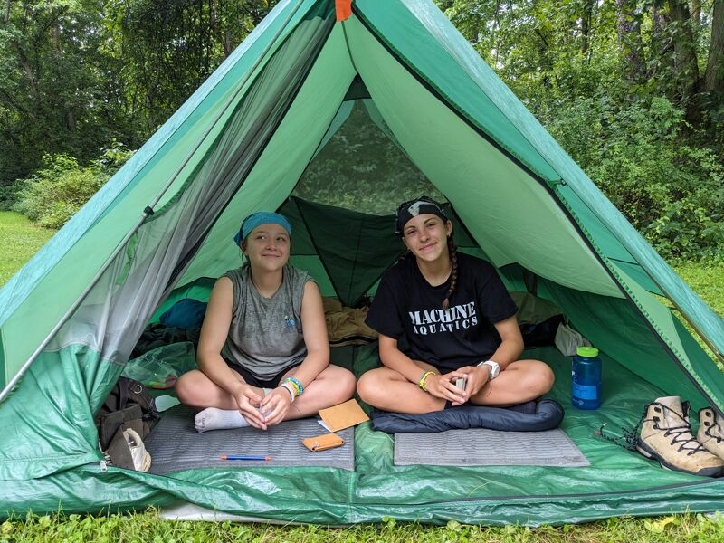 Two young women are sitting cross-legged inside a green tent. They are both smiling and appear to be enjoying their camping trip. The tent is set up on a grassy area, and there are trees in the background. One woman is wearing a blue headscarf and a gray tank top, while the other is wearing a black headband and a black t-shirt. There are camping supplies scattered around them.