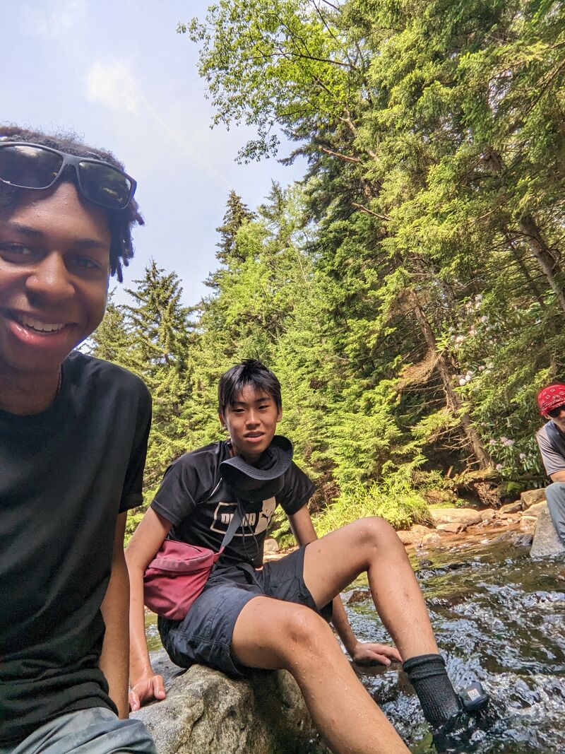 The image shows three young people in a natural setting, possibly a forest or park. Two of them are prominently featured in the foreground, with one wearing sunglasses and a black t-shirt, and the other sitting on a rock near a stream. The third person is partially visible in the background. The environment includes lush greenery and a flowing stream, suggesting an outdoor adventure or leisurely activity.