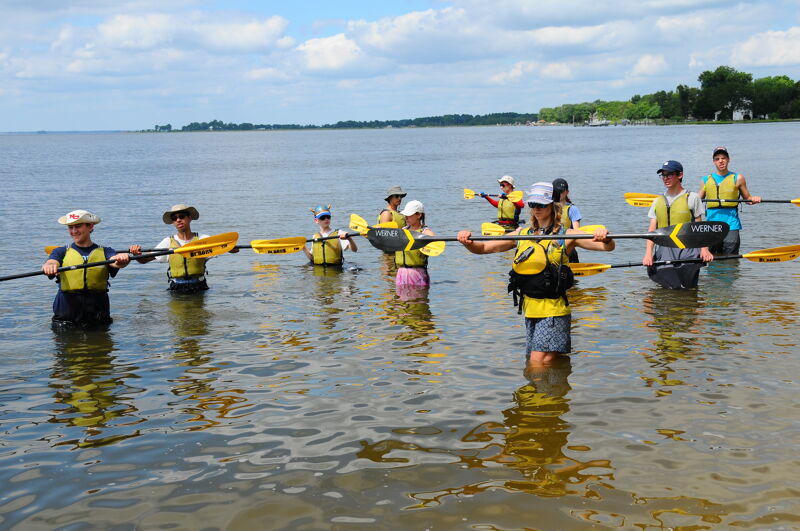 A group of people are standing in shallow water, holding paddles. They appear to be preparing for or just finishing a kayaking or canoeing activity. The water is calm, and the sky is partly cloudy. There's land visible in the background, suggesting they are near a shore.