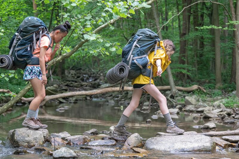 Two young women are carefully crossing a shallow stream by stepping on rocks. They are both wearing large backpacks and hiking attire, suggesting they are on a backpacking trip. The surrounding environment is a lush green forest, with trees lining the stream. The sunlight filters through the leaves, creating a dappled effect on the water and rocks.