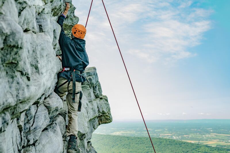 A person wearing a helmet is rock climbing on a steep rock face. They are using ropes and gear for safety. The background shows a scenic view with a blue sky, some clouds, and a distant landscape with trees and possibly a body of water. The climber is focused and determined as they ascend the rock.