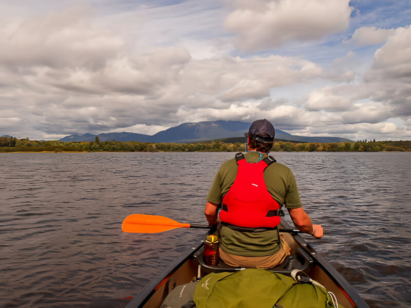 A person wearing a red life jacket and a baseball cap is paddling a canoe on a lake. The person is facing away from the viewer, and the canoe is in the middle of the frame. The lake is surrounded by trees and mountains in the distance. The sky is cloudy.