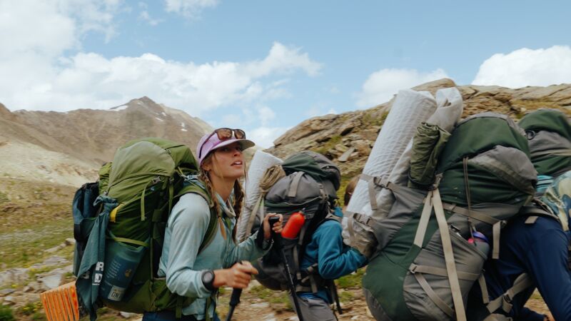The image shows three people hiking in a mountainous region. They are carrying large backpacks, indicating they are likely on a multi-day trek. The sky is partly cloudy, and the landscape appears rugged and natural. The hikers are dressed in outdoor gear, prepared for the elements. The overall impression is one of adventure and exploration in a remote, scenic environment.