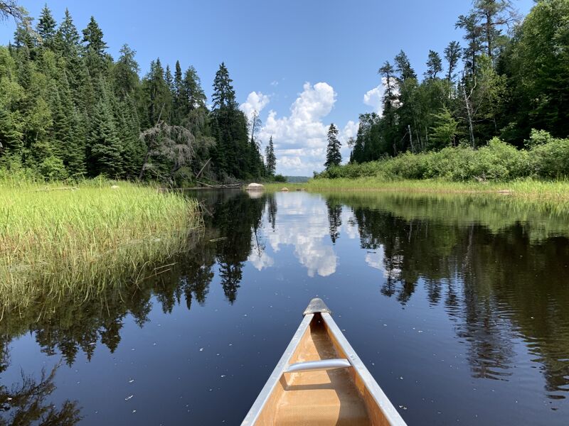 A canoe glides through calm waters, reflecting the sky and surrounding lush greenery. Tall trees line the riverbanks, creating a serene and picturesque scene. The clear water mirrors the clouds above, enhancing the tranquil atmosphere. The canoe's wooden frame adds a touch of rustic charm to the natural beauty of the landscape.