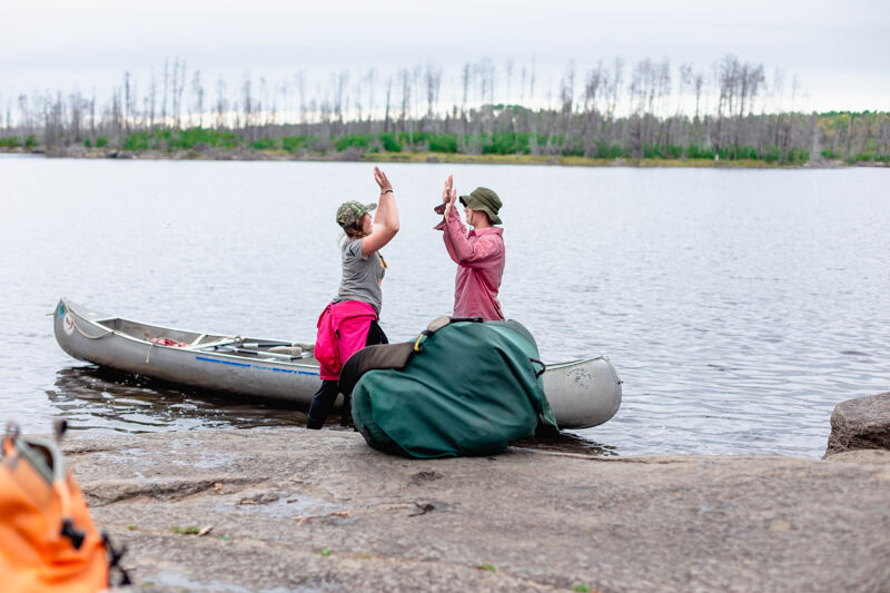 Two people are giving each other a high five next to a canoe on the shore of a lake. One person is wearing a pink skirt and a hat, and the other is wearing a red shirt and a hat. There is a large green bag next to the canoe. The lake is calm and there are trees in the background.