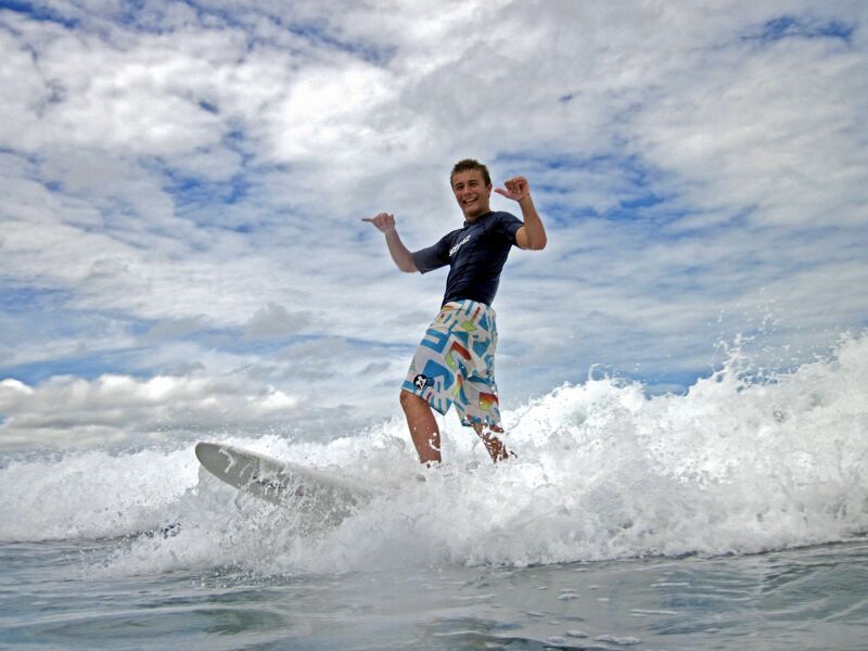 A surfer is riding a wave. He is standing on the surfboard with his arms raised in the air, appearing joyful and triumphant. The wave is breaking, creating white foam around the board. The sky is blue with fluffy white clouds, suggesting a sunny day. The surfer is wearing a dark top and patterned shorts, indicating he is dressed for the water sport.