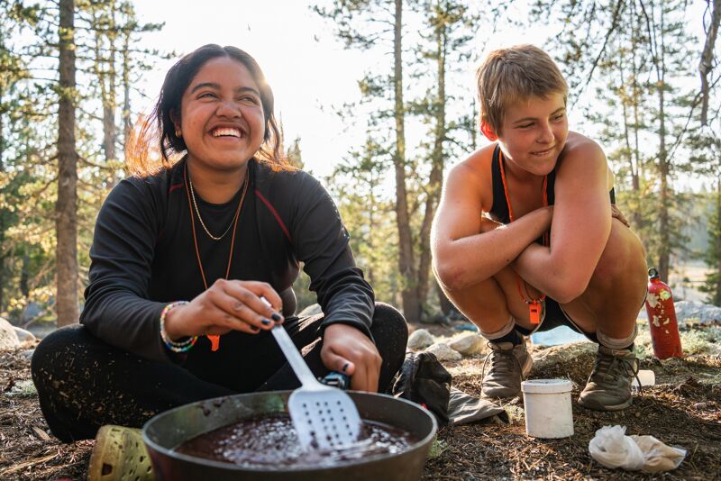 Two young women are outdoors, possibly camping. One is sitting and stirring something in a pot with a spatula, smiling. The other is crouched nearby with her arms crossed, also smiling. They appear to be preparing food. The background shows trees and a natural setting, suggesting they are in a forest or wilderness area.