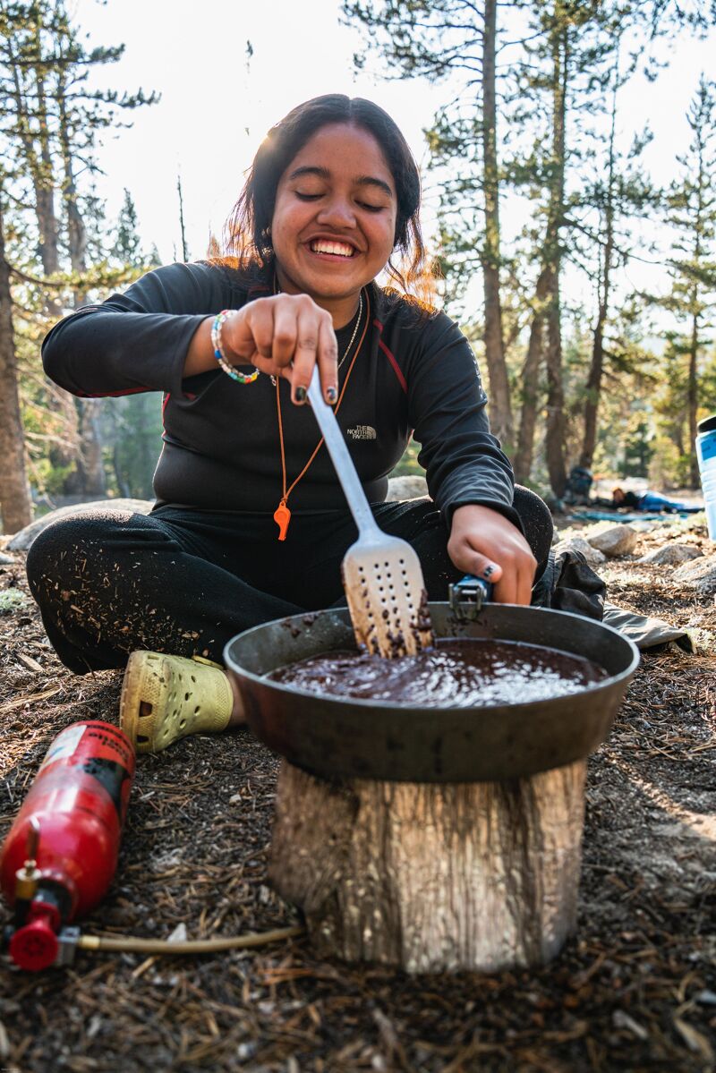 A woman is cooking outdoors, smiling as she stirs a pot of food over a portable stove. She's wearing a dark long-sleeved shirt and dark pants, suggesting she's camping or hiking. The cooking setup is on the ground, with a red propane tank nearby. The background shows a forest setting with trees and sunlight filtering through the leaves.