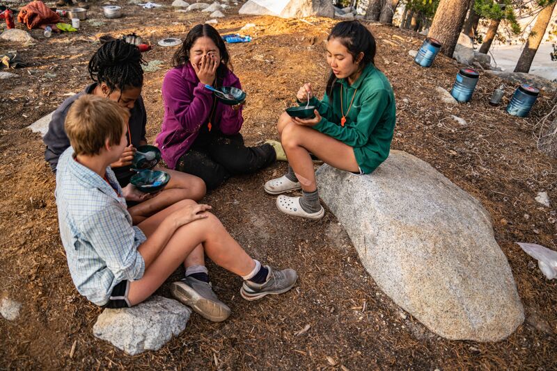 The image shows a group of four young people sitting outdoors, possibly camping. They appear to be eating from small bowls. One person is sitting on a large rock, while the others are on the ground or smaller rocks. The setting looks like a forest or mountainous area, with trees and rocky ground visible.