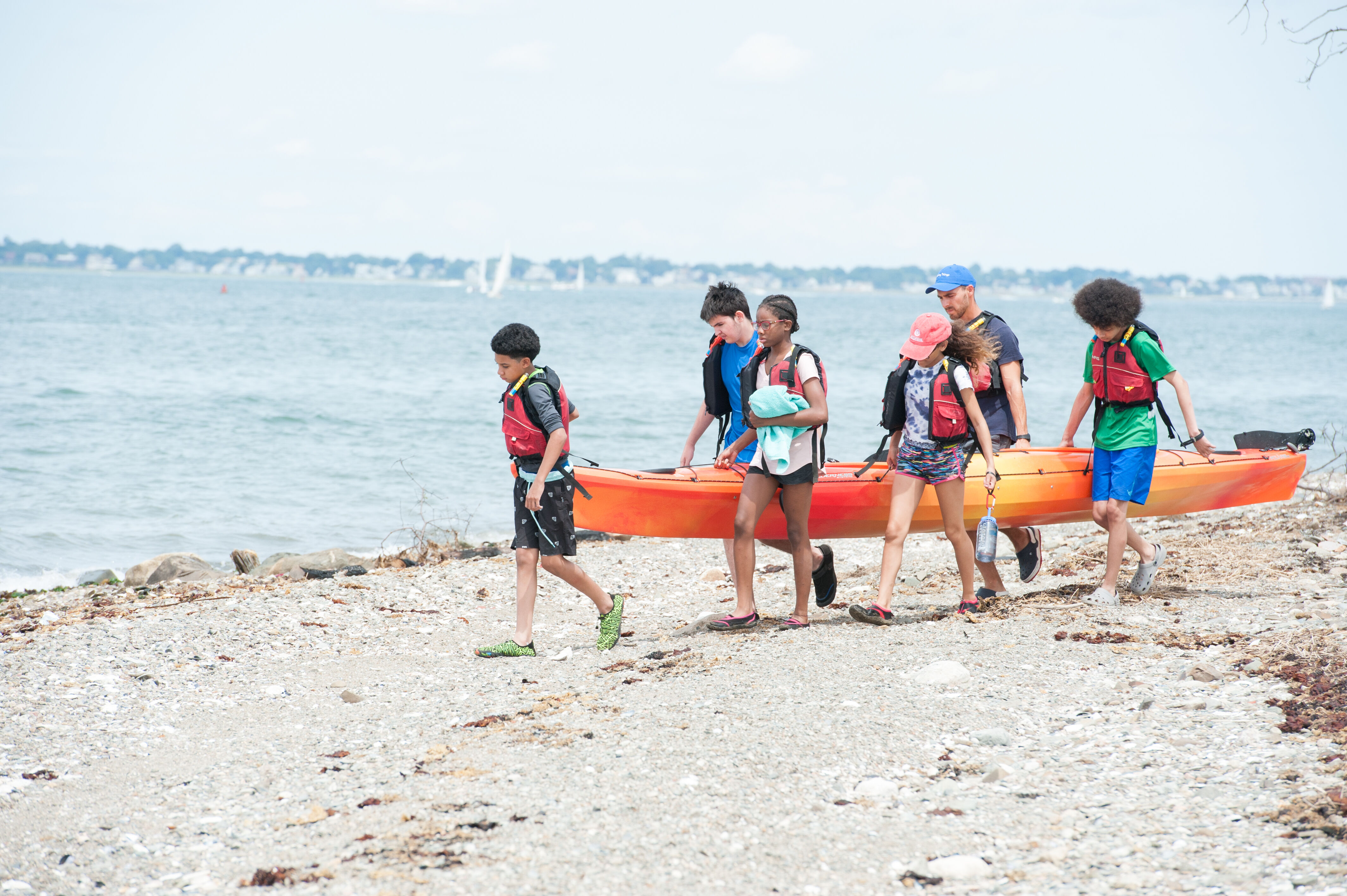 In the image, a group of people is carrying a kayak on a beach. They are all wearing life jackets and appear to be heading towards the water. The sky is overcast, and the sea is calm. The scene suggests a day of water activities.