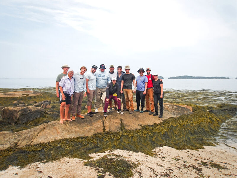 A group of approximately 15 people are standing on a rocky, seaweed-covered shore near the ocean. They are casually dressed and appear to be posing for a photo. The sky is overcast, and there's an island visible in the distance. The overall scene suggests a coastal or island setting, possibly during a group outing or excursion.