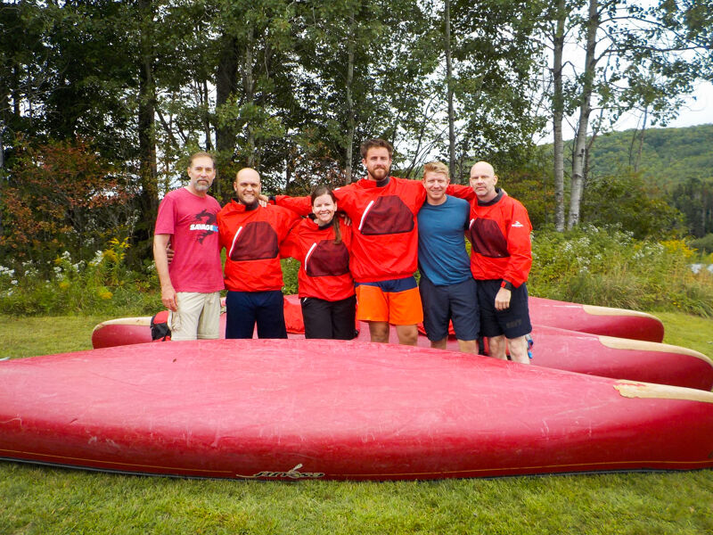 A group of seven people stands behind a stack of red canoes on a grassy field. They are all wearing red jackets, with one person in a pink shirt. The background features trees and a hill. The photo appears to be taken outdoors on a bright day.