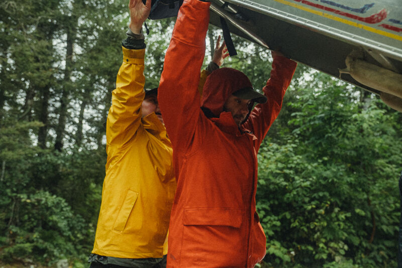 Two people in rain gear lift a canoe over their heads in a forest setting. The person in the foreground wears a bright orange jacket with the hood up, while the person behind them is wearing a yellow jacket. The trees in the background are lush and green, suggesting a natural environment. The lighting indicates it might be an overcast or rainy day.