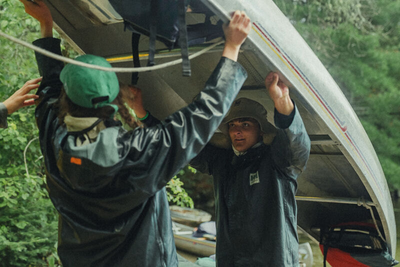 Two people are lifting a canoe. The person on the left is wearing a green hat and a black jacket. The person on the right is wearing a hat and a black jacket. They appear to be preparing for a canoeing trip, possibly near a wooded area. The canoe is silver and seems lightweight, suitable for carrying. Other canoes are stacked in the background.