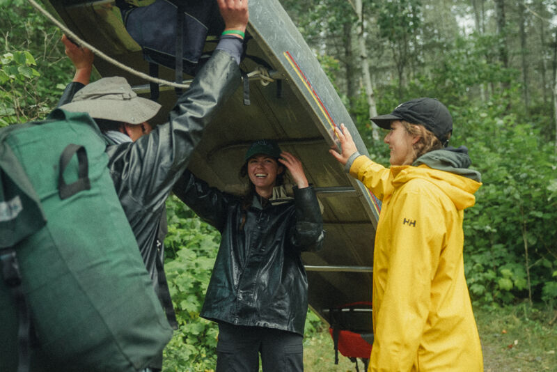 Three people are carrying a canoe through a wooded area. The person on the left has a large green backpack and is wearing a hat and a dark raincoat. The person in the middle is under the canoe, wearing a black raincoat and smiling. The person on the right is wearing a yellow raincoat and a black baseball cap. The trees in the background suggest they are in a forest or park.
