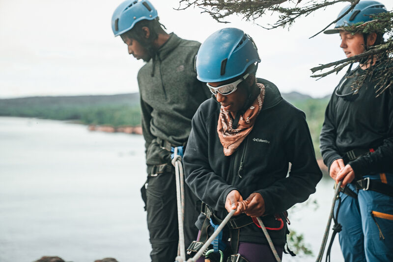 The image shows three people wearing helmets and harnesses, likely preparing for or engaging in a climbing or rappelling activity. They are standing near a body of water, possibly a lake or river, with a forested area in the background. The person in the center is looking down and appears to be adjusting their equipment, while the other two are looking in different directions. The overall scene suggests an outdoor adventure or recreational activity.