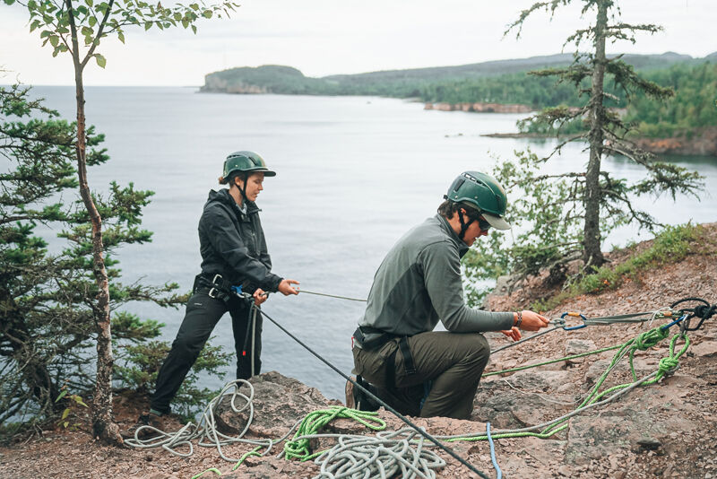 Two people are on a rocky cliff overlooking a body of water. The person on the left is standing and holding a rope, while the person on the right is kneeling and adjusting the rope. Both are wearing helmets and harnesses, suggesting they are engaged in some kind of climbing or rappelling activity. The background features a scenic view of the water, trees, and distant land.
