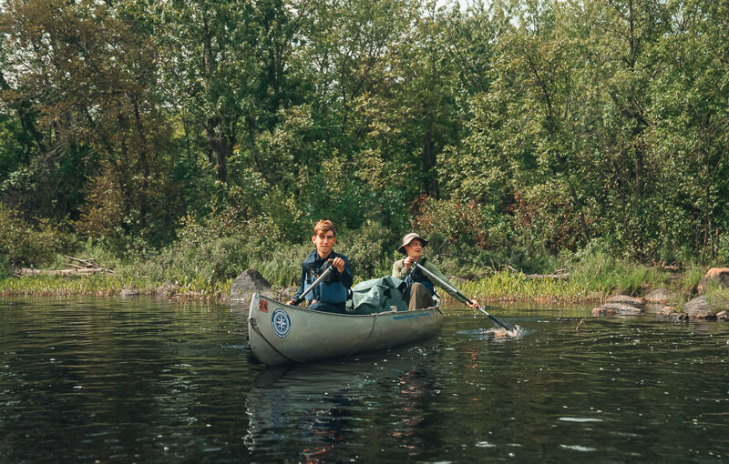 Two people are paddling a canoe on a calm body of water, likely a lake or river. The canoe is in the center of the frame, with the paddlers positioned at the front and back. Lush green trees and vegetation line the shore in the background, creating a natural and serene setting. The water reflects the surrounding greenery, adding to the peaceful atmosphere of the scene.
