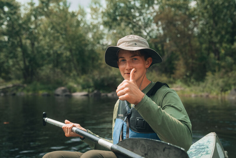 A young person wearing a hat and a green long-sleeve shirt is sitting in a canoe on a lake or river. They are holding a paddle and giving a thumbs-up. The background shows a lush green forest. The water is calm, and the overall scene suggests a peaceful outdoor activity like canoeing or kayaking. The person appears to be enjoying their time on the water.