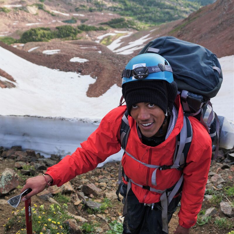 A man is hiking up a mountain. He is wearing a red jacket, a blue helmet, and a large backpack. He is using an ice ax to help him climb. There is snow on the ground and the sky is overcast. The man is smiling and looks like he is enjoying his hike.