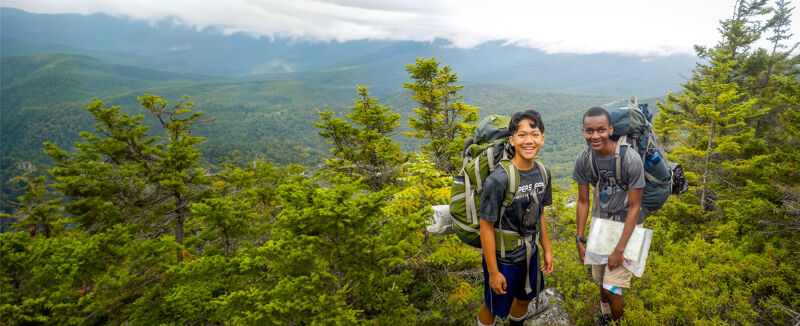 Two young hikers are seen on a mountain trail, surrounded by lush green trees and a scenic view of distant mountains. Both are wearing backpacks, and one is holding a map. They appear to be enjoying their outdoor adventure, pausing to take in the beautiful natural surroundings. The sky is overcast, creating soft lighting for the scene.