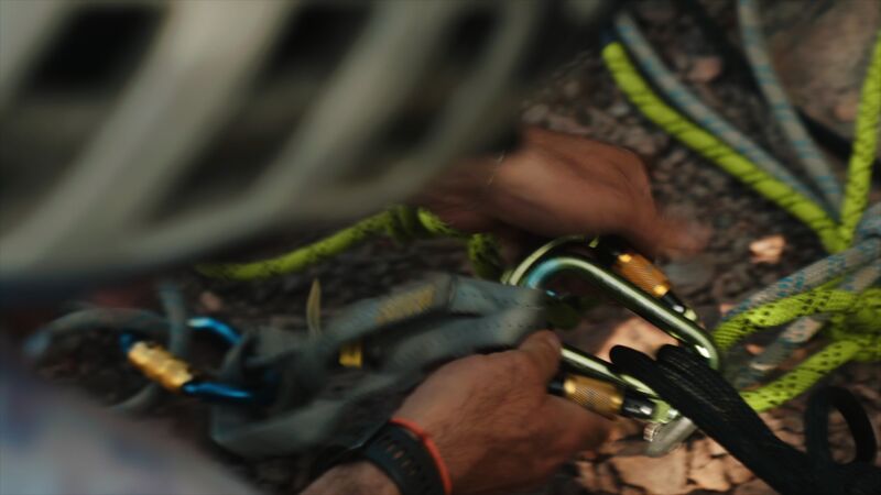 From a high-angle perspective, the image captures a climber's hands meticulously organizing climbing gear. Carabiners, ropes of vibrant green and blue hues, and a grey gear sling are arranged on a rocky surface. The climber, wearing a helmet and a watch, is focused on preparing the equipment, suggesting a moment of concentration before or after a climb. The composition emphasizes the importance of preparation and safety in the sport of climbing.