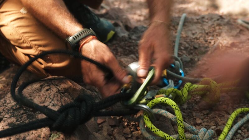 A close-up shot shows a person's hands tying a knot with climbing ropes. The ropes are of different colors, including black, green, and yellow. The person is wearing a watch and a bracelet. The background is blurred, but it appears to be a rocky outdoor setting. The focus is on the intricate knot-tying process, highlighting the skill and precision required for climbing activities.