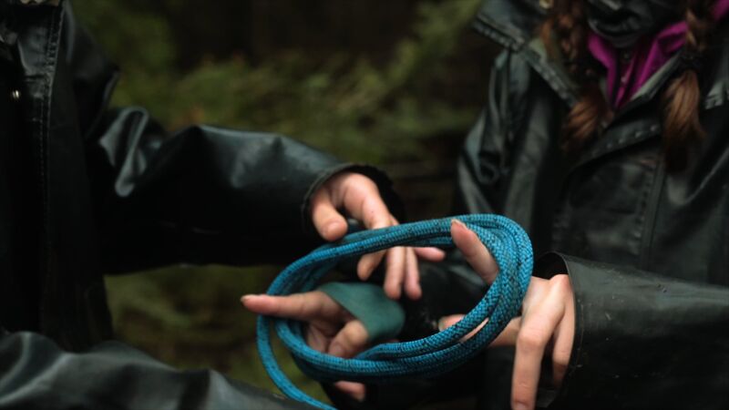The image shows two people holding a blue rope. The person on the left is holding the rope with their left hand, while the person on the right is holding the rope with their right hand. Both people are wearing black jackets. The background is blurry, but it appears to be a forest.