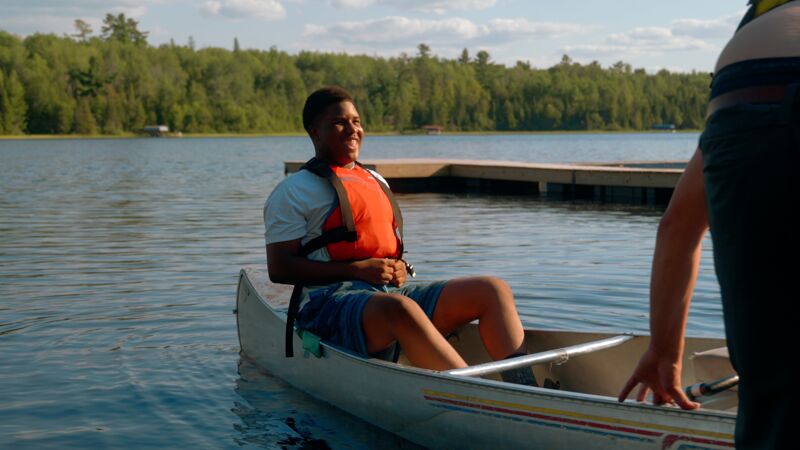 A young man wearing a life vest is sitting in a canoe on a lake. He is smiling and appears to be enjoying himself. In the background, there is a wooden dock and a forest. Another person is partially visible on the right side of the image, near the canoe.