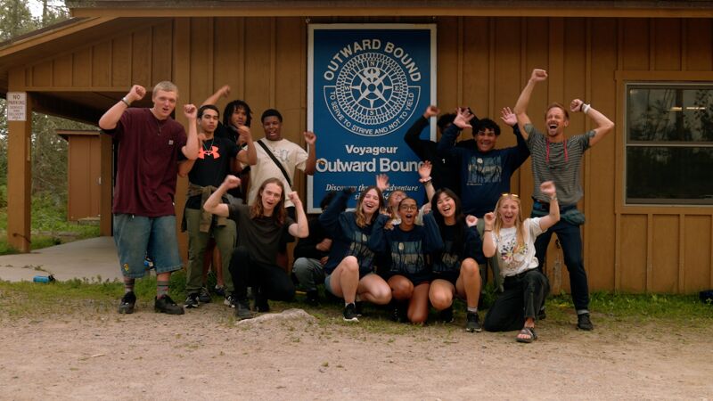 A group of people are posing for a photo in front of a building with an "Outward Bound" sign. They are all smiling and have their arms raised in the air, appearing excited and cheerful. The group consists of both men and women of varying ages. The building is a light brown color, and the photo seems to be taken outdoors during the daytime.